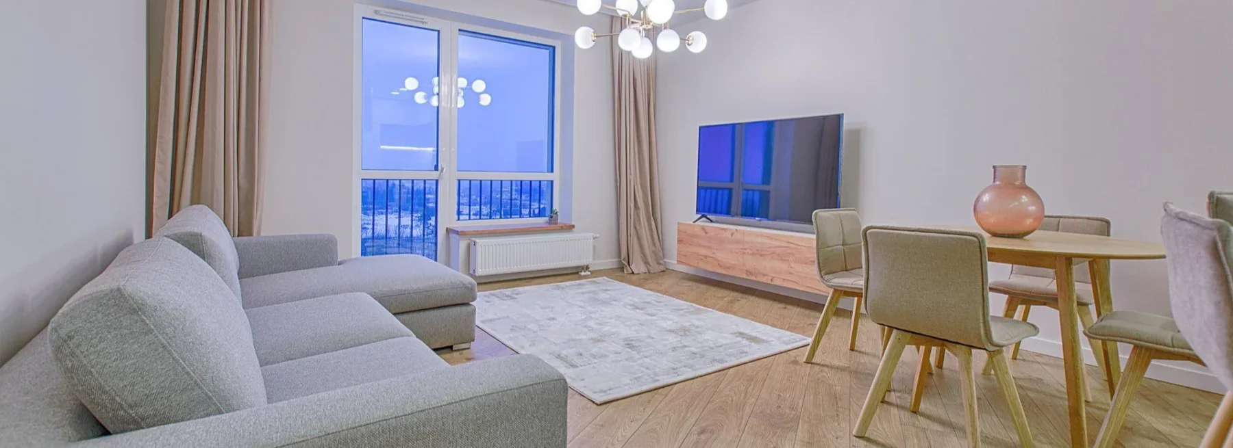 Modern living room with gray sofa, dining table, TV, and floor-to-ceiling windows overlooking city at dusk