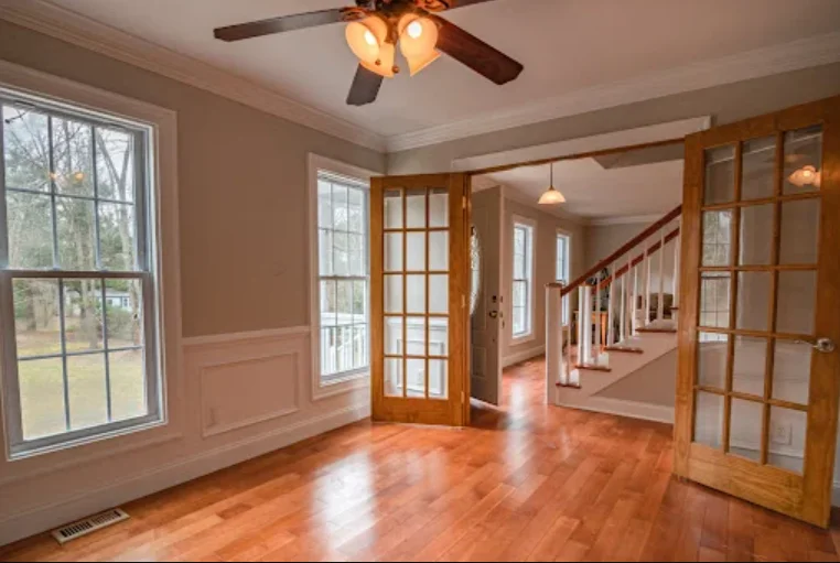 Spacious empty room with hardwood floors, French doors, large windows, ceiling fan, and staircase